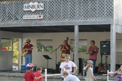 Outdoor stage with musicians performing; three standing with instruments and one seated performer, audience watching; MINBURN banner overhead.